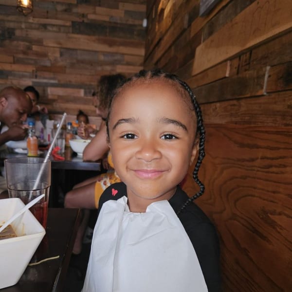 A smiling child wearing a bib at Roc N Ramen, with other diners in the background.