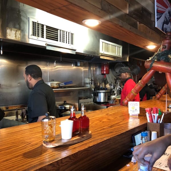 Two Roc N Ramen chefs work in a stainless steel kitchen behind a wooden counter with condiments.