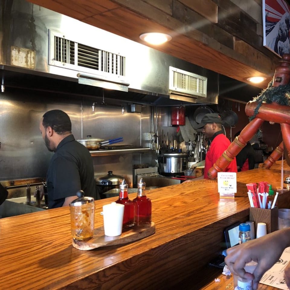 Two Roc N Ramen chefs work in a stainless steel kitchen behind a wooden counter with condiments.