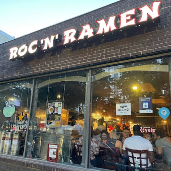 Exterior of Roc N Ramen restaurant with a large sign, visible diners, and a "Now Hiring" sign.