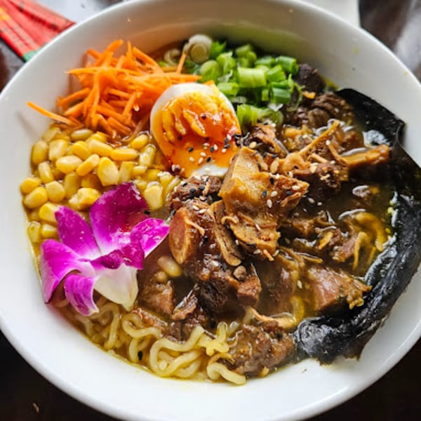 A bowl of Roc N Ramen with noodles, meat, corn, carrots, egg, green onions, and a purple orchid.